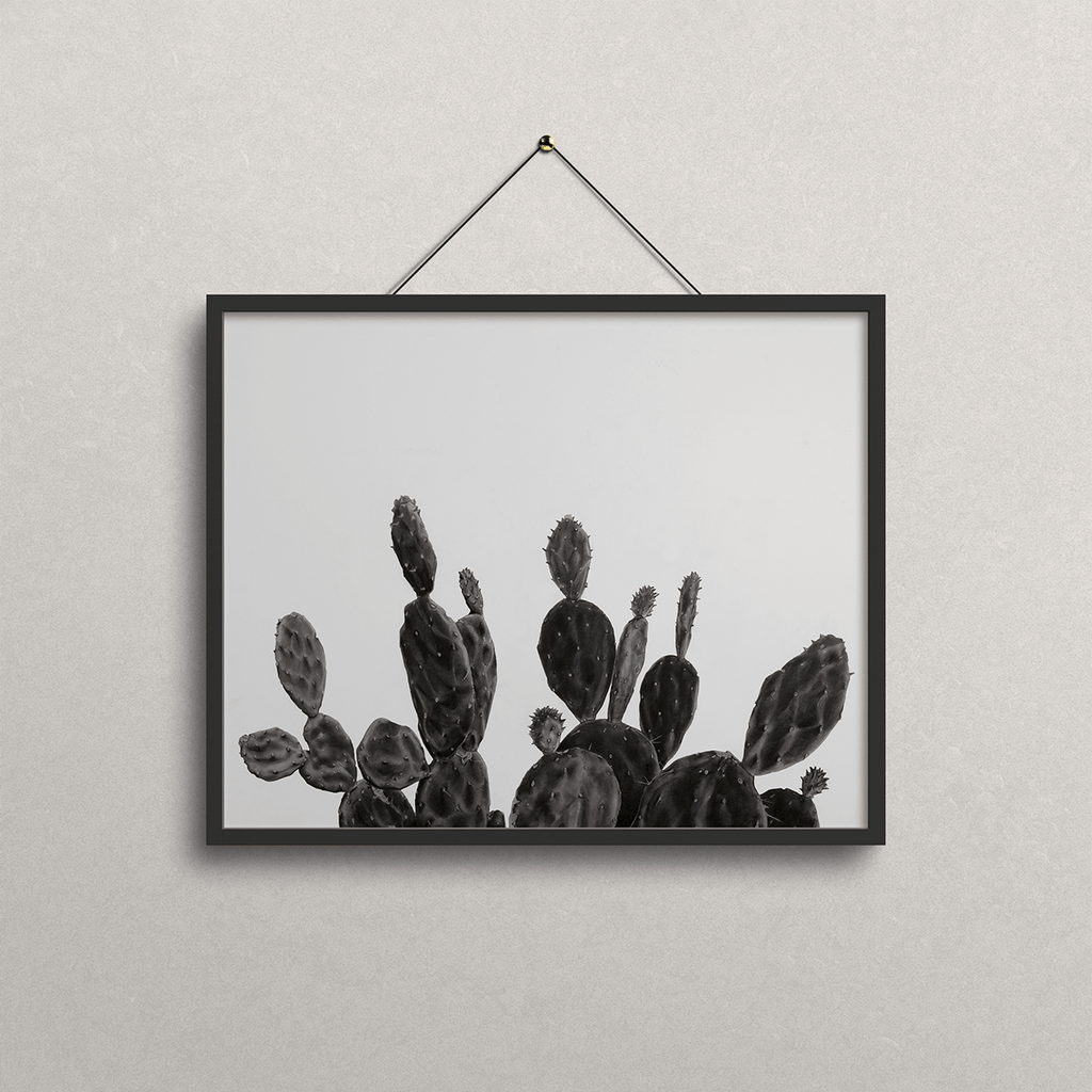 A black-framed wall art print featuring a black and white drawing of prickly pear cacti, hanging from a black cord on a textured light gray wall.