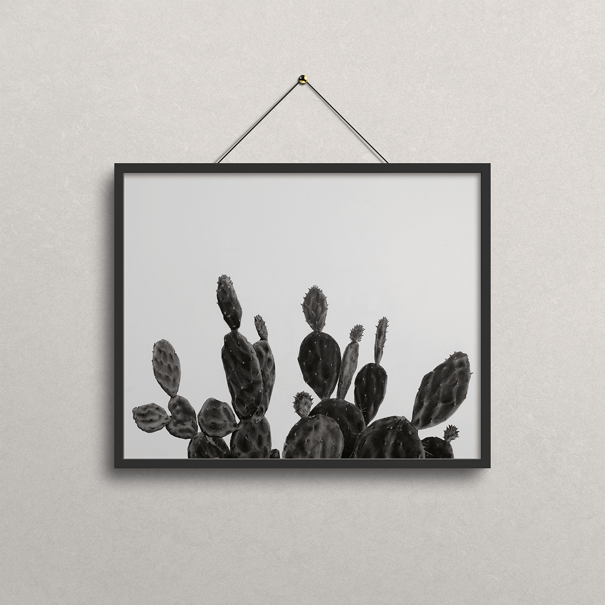 A black-framed wall art print featuring a black and white drawing of prickly pear cacti, hanging from a black cord on a textured light gray wall.