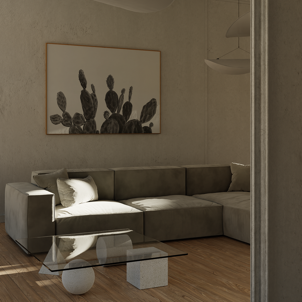 A modern living room scene featuring a low gray sectional sofa with a large framed black and white cactus drawing on the textured beige wall above it. A glass-top coffee table sits in front of the sofa on light wood flooring.
