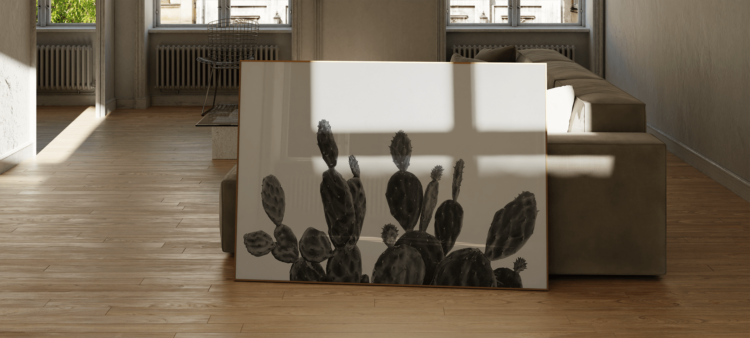 A large art print featuring a black and white illustration of prickly pear cacti, leaning against a beige sofa in a modern, sunlit living room with light wood floors.