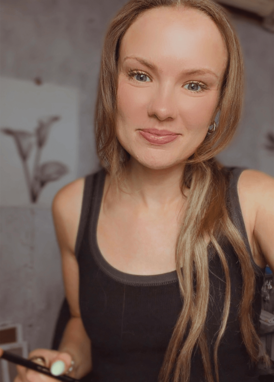 A woman with light brown hair wearing a black tank top, holding a charcoal pencil in a softly lit indoor setting.