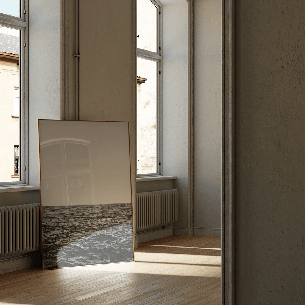 A large framed art print leaning against a window in a sunlit room with wood floors and cream walls.