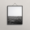 A black-framed wall art print hanging on a light gray textured wall. The print shows a black-and-white seascape photorealistic drawing with a pale sky above dark ocean water.