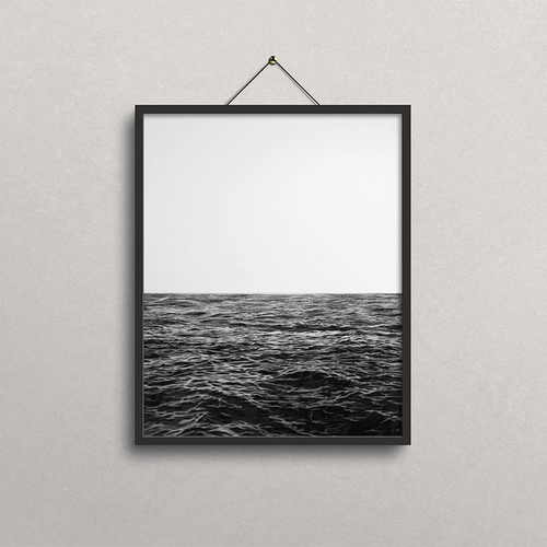 A black-framed wall art print hanging on a light gray textured wall. The print shows a black-and-white seascape photorealistic drawing with a pale sky above dark ocean water.