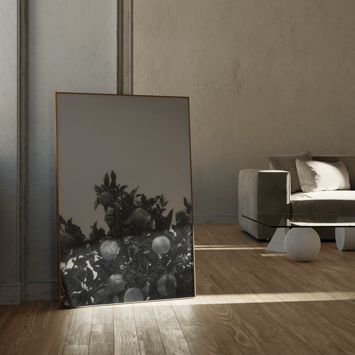 A large vertical framed art print featuring a black and white drawing of an orange tree, leaning against a textured wall in a modern, minimalist living room with a gray sofa and light wood flooring.