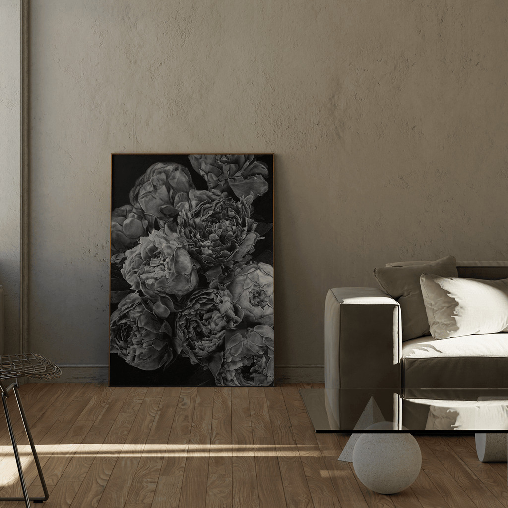 A vertical black and white floral art print depicting peonies, displayed in a modern living room setting next to a gray sofa and wooden floor.