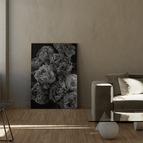 A vertical black and white floral art print depicting peonies, displayed in a modern living room setting next to a gray sofa and wooden floor.